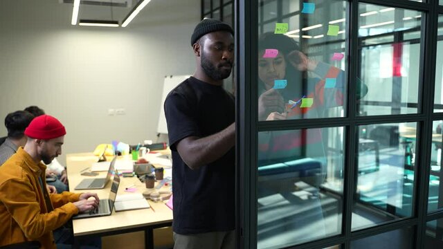 Focused dark-skinned man and woman workin on sticky notes on joint student's project. Young people standing at glass partition with sticky notes while other classmates working at table