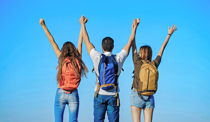 Friends with backpacks outdoor on sunny day with raised hands on blue sky background. Carefree happiness freedom concept. Tourists on nature.