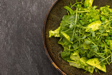 Green vegetables salad sweet pea, avocado, arugula, cucumber in a black bowl on a dark slate, stone or concrete background.Top view.