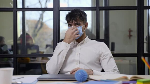 Close-up portrait of confident handsome hindu man putting on protective mask while posing on camera at workstation. Masked young freelancer sitting at desk in contemporary office with glass partitions