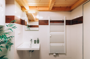 Bathroom wall with white washbasin and chrome faucet. On the wall there is mirror with bright glowing light and next to it is heating ladder. On the left is partly visible boiler and decorative plant.