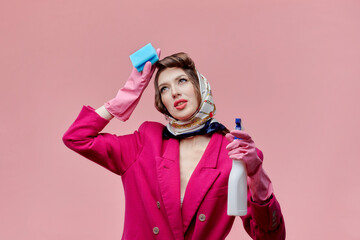 Young woman tired after cleaning the house, hand near her forehead. In the hands of spray and sponge. Bright makeup. Pin-up style. Pink background.