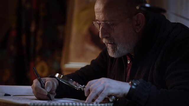 Close-up of an artist working in the studio. Older man painting a picture with a magnifying glass in a creative environment.