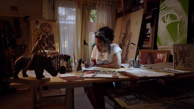 Camera zooming in on a young artist drawing a portrait of a man sitting at the studio work table. African American clothing designer designing new men's collection.