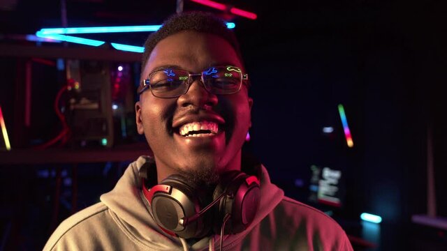 A kind dark-skinned guy with glasses laughs, looking at the camera, with headphones around his neck