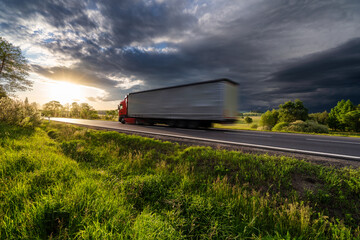 Motion blurred red truck driving on the asphalt road in rural landscape at sunset with dark storm...