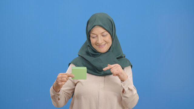 The old woman in a turban holding a green box in her hand points to the box with her finger and makes a thumbs up. Studio shot isolated on blue background.