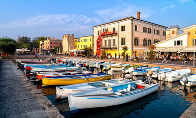 Fototapeta na wymiar Bardolino, Italy, 10/28/2019: Boats in old town port of Bardolino and tourists walking and sitting in restaurants. The town is a popular holiday destination in Garda Lake district.