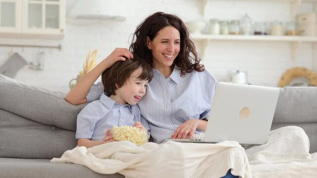 Smiling mother with little preschool son watch funny video, cartoon on laptop computer sit together on sofa in living room. Young mom and cute boy with popcorn under blanket spend time at funny movie