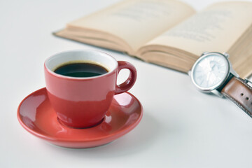 Black coffee in red cup with wrist watch and book is blurred background.