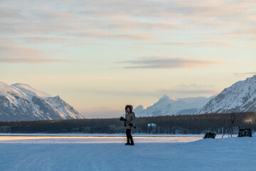 Woman holding a tripod and camera walking along a frozen lake in northern Canada on a cold, sunset afternoon in spring time April. Wearing beige jacket and black pants. 