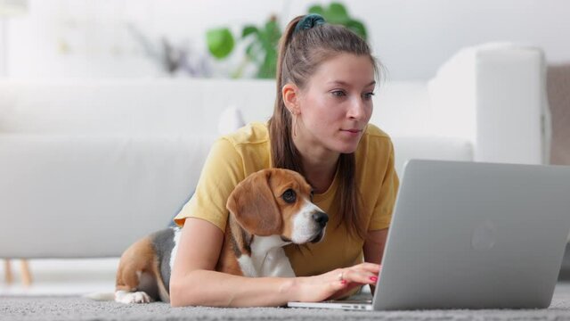 Woman student lying on floor with laptop and doing typing homework task with pet dog Spbi. 20s