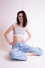 woman in blue jeans and sports top sits, white studio background