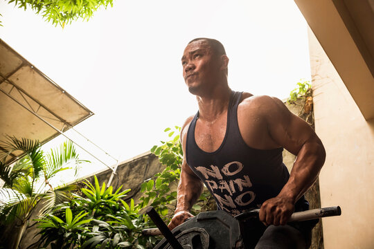 A brawny and well muscled man does t-bar rows at his backyard. Working out and weight training at home.
