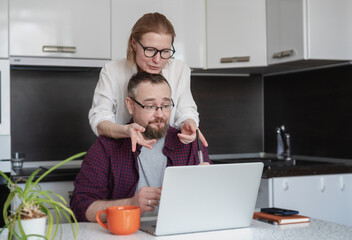 Young adult couple husband and wife tsitting in the kitchen and looking at a laptop screen., checking banking online application on a laptop