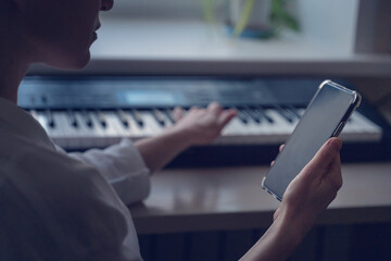 Adult young blonde woman teacher playing piano and checking notes on smartphone, self-education music app