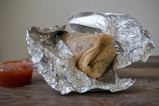 A Whole Carne Asada Burrito Wrapped In Open Foil Sitting On A Wood Table.