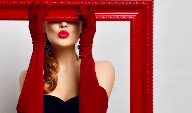 Portrait of excited young woman artist in black off-shoulder tight dress, red elbow-length gloves holding big red empty picture frame covering her eyes and sending, blowing kiss