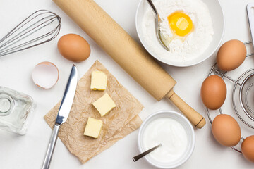 Brown eggs on metal stand. Butter and knife on paper. Egg yolk with flour and spoon in bowl. Milk in bowl and rolling pin on table