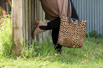 Feet only fashion image of woman wearing leopard skin boots with bag leaning against wooden fence. 