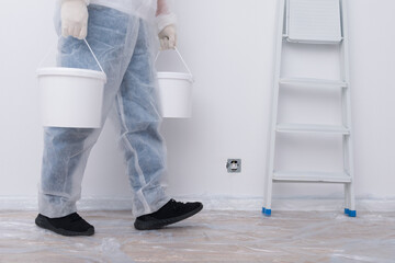 a man in protective clothing, bringing buckets of paint, against the background of a white wall and a stepladder © kurgu128