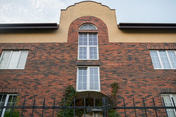 View from behind black wrought-iron fence on facade of two-story town house lined with decorative bricks with arched top of middle part with high windows and entrance covered with green wild grapes.