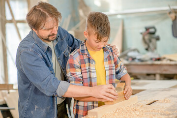 Father teaches to young boy to plan wood in a carpentry workshop. Hobby and education concept