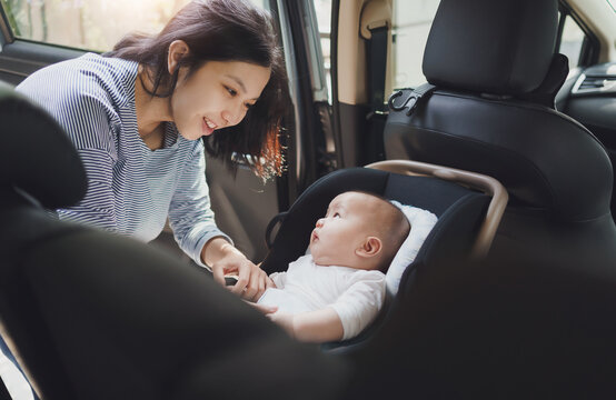 Happy Smiling Beautiful Asian Young Mother Putting Her Baby Son Into Car Seat And Fasten Seat Belts In The Car