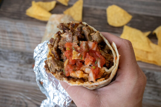 A hand holding a carne asada grilled steak burrito with salsa over a table with tortilla chips