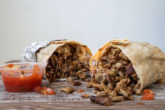 A carne asada burrito cut in half with meat falling out onto the table next to a cup of salsa.