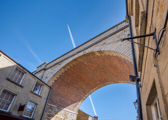 Jumbo jet aeroplane flying over railway bridge arch viaduct in blue sky with vapour trails. Low angle aircraft high in the sky looking up under old historical red brick archway