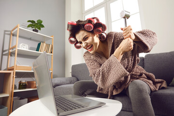 Funny furious woman in hair curlers breaking and destroying laptop computer with hammer. Angry...