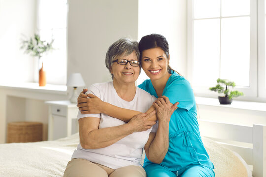 Portrait Of Supportive Carer With Her Elderly Patient Who Has Alzheimer. Happy Retired Senior Woman Together With Caring Young Nurse Or Caregiver Hugging And Smiling Sitting On Bed In Retirement Home