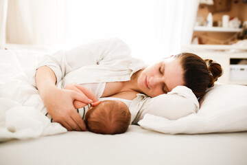 Obraz na płótnie Canvas Cute caucasian mom and newborn baby, mom breastfeeds her baby lying on a white background on the bed in the bedroom, close-up, soft focus