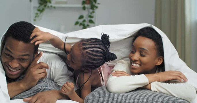 African family lies under blanket, little daughter active child touches pokes finger in male eye of dad black man father suffering from allergy pain of poor vision, afro parents with baby kid portrait