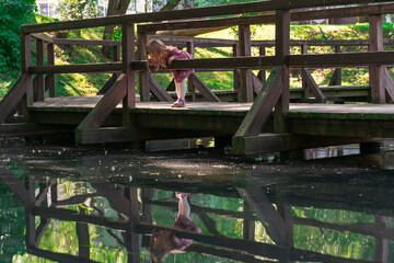 little girl in a red dress looks at her reflection in a pond standing on a wooden bridge