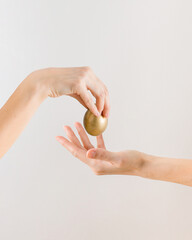 Beautiful woman's hand puts golden Easter egg in the other hand. Minimal creative Easter gift concept on the pastel beige background with a copy space.