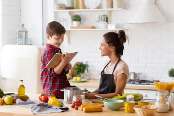 Happily smiling mother and son preparing dinner on loft kitchen. Boy sitting on table reading recipe from cookbook to cheerful mom wearing apron. Happy family weekend. Excited housework