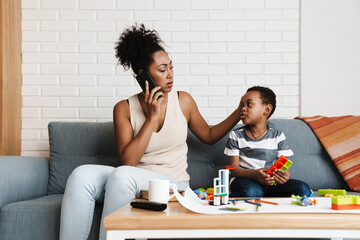 Black woman talking on cellphone while her son playing with toys at home