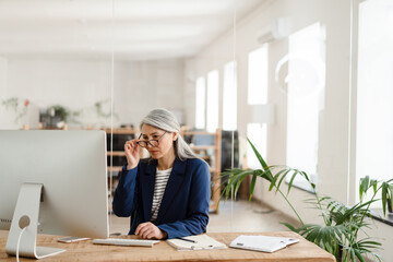 The Asian woman sitting at a table in front of the monitor adjusting glasses with her hand in the office