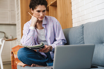 A woman sitting in front of a laptop and writing something with her hand on her chin in the room