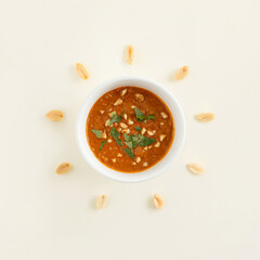 Peanut kernels arranged in a circle around the bowl with African piquant delicious spicy peanut coconut sauce on pastel beige background. Minimal organic natural healthy food flat lay summer concept.