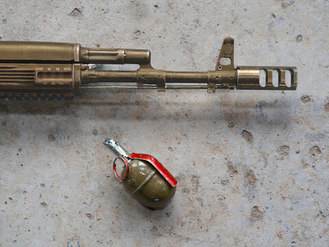 An assault rifle and a hand-held offensive grenade lie in close-up on a gray concrete floor view from above. Gold skin for weapons in a tactical kit. The concept of modern therorism and game cosplay.