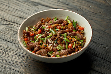 Delicious Asian street food - wok rice with beef, green onions, vegetables and sesame seeds in a white bowl on a wooden background.