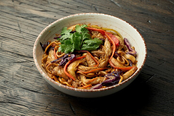 Delicious asian street food - egg noodles with chicken, cilantro, vegetables and scrambled eggs in a white bowl on a wooden background. Wok noodles