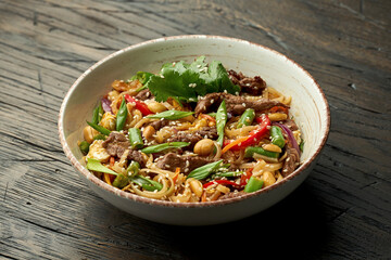 Delicious asian street food - pad thai noodles with beef, cilantro, vegetables and scrambled eggs in a white bowl on a wooden background.