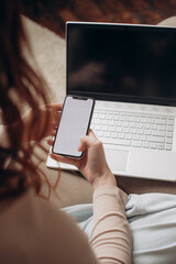 a girl holds a smartphone with a blank screen in her hands and runs her finger across the screen, against the background of a laptop, diary and tea