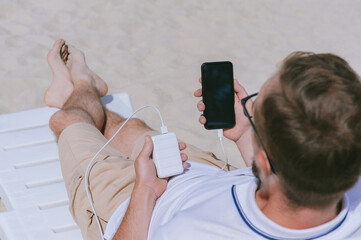A man lies on a sun lounger with a mock-up of a smartphone and is charging from a Power Bank. Against the background of sand.