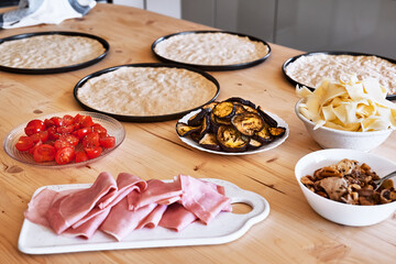 Ingredients for homemade Italian pizza. Pizza dough, tomato, cheese, eggplant, mushroom and ham in plates on the table in the kitchen. Selective focus