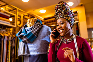 tanzanian woman with snake print turban over hear working in fabrics shop calling to client by smartphone - obrazy, fototapety, plakaty